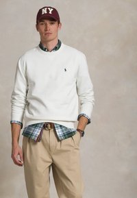 Maglione bianco con logo blu, indossato sopra una camicia a scacchi verde e blu, abbinato a pantaloni beige e un berretto maroon di New York.