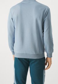 Sudadera azul claro con cuello y puños acanalados, que presenta una textura suave y un corte holgado. Combinada con pantalones oscuros.