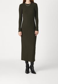 Dlouhá žebrovaná šaty v tmavě zelené barvě s dlouhými rukávy a kulatým výstřihem, doplněné černými kozačkami po kolena a stříbrným řetízkovým náhrdelníkem.