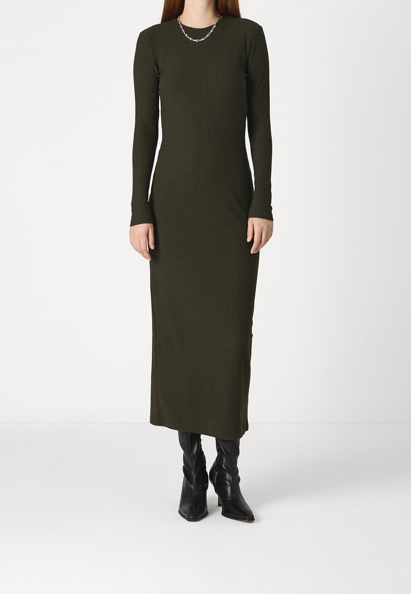 Dlouhá žebrovaná šaty v tmavě zelené barvě s dlouhými rukávy a kulatým výstřihem, doplněné černými kozačkami po kolena a stříbrným řetízkovým náhrdelníkem.