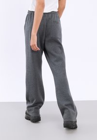 Person som bär löst sittande mörkgrå sweatpants med bakfickor, en vit randig ärmlös topp och svarta chunky skor, stående på en grå golv.