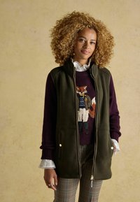 Olivovo zelená vestu na zips over burgundy sveter s dizajnom líšky, s bielym riaseným golierom a kockovanými nohavicami. Zlaté doplnky.