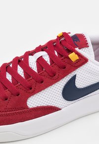 Röd mocka och vit nät-sneaker med marinblå Nike-swoosh, röda snören och gula accenter på snörena, med en låg profil design.