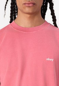 Pink cotton t-shirt with a crew neckline and short sleeves, featuring a small white "obey" logo on the chest. Soft texture.