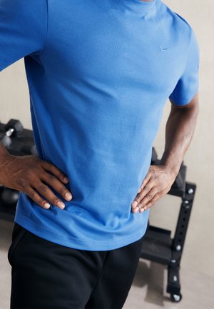 Osoba w niebieskiej sportowej koszulce i czarnych szortach stoi z rękami na biodrach obok stojaka z hantlami na siłowni.