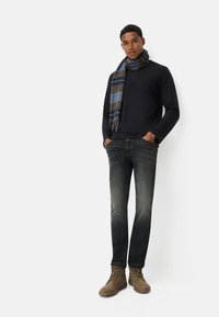 Chemise à manches longues bleu marine foncé avec trois boutons, associée à un jean foncé déchiré. Les accessoires incluent une écharpe rayée marron et bleu, ainsi que des bottes kaki.