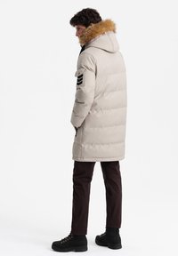 Giacca a vento leggera beige, con cappuccio foderato in pelliccia, cerniere laterali e una toppa sul braccio. Indossata con pantaloni marroni e stivali neri.