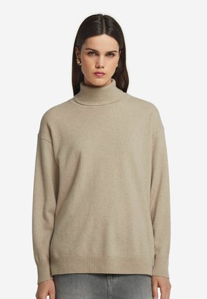 Frau mit langen, dunklen Haaren, die goldene Hoops-Ohringe, einen beigen Rollkragenpullover und graue Hosen trägt, vor einem schlichten weißen Hintergrund.