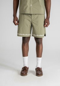 Oliven grønne shorts med lodrette striber, blomsterbroderi ved kanten, paret med hvide sokker og brune snørebåndssko.