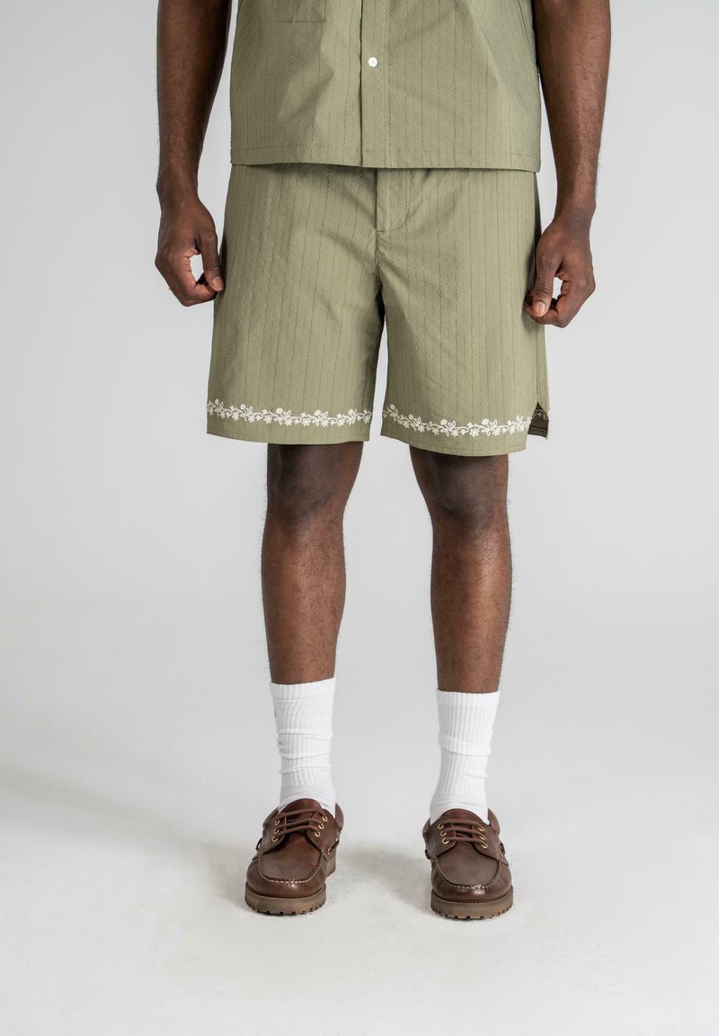Oliven grønne shorts med lodrette striber, blomsterbroderi ved kanten, paret med hvide sokker og brune snørebåndssko.