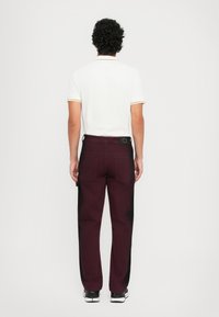 Tenue masculine comprenant un polo blanc avec des accents jaunes et un pantalon bordeaux. Le pantalon a une ceinture noire et une poche latérale.