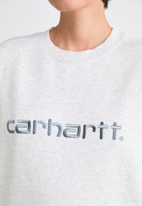 Lichtgrijze sweatshirt met een geribde ronde halslijn en een groot blauw geborduurd "carhartt"-logo over de borst. Zachte stoftextuur.