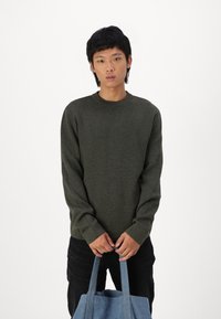 Mørkegrønn genser med lange ermer, rund hals, teksturert stoff. Person som holder en lyseblå totebag. Enkel, uformell design.