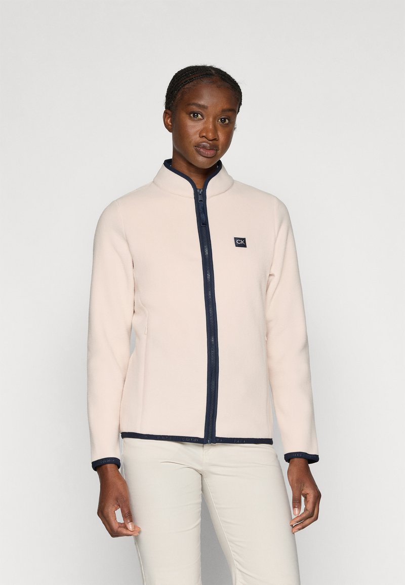 Hellrosa Fleecejacke mit hohem Kragen, marineblauem Reißverschluss und Besatz. Verfügt über seitliche Taschen und ein kleines Logo auf der Brust.