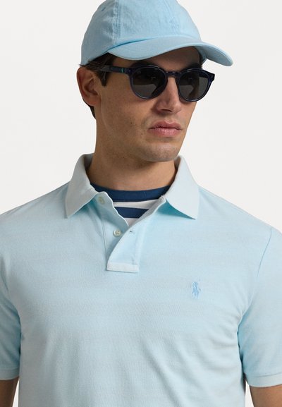 Polo shirt de culoare albastră deschis, cu un material texturat și guler cu nasturi, purtat peste o cămașă în dungi albă și bleumarin. Asortat cu o șapcă potrivită și ochelari de soare.