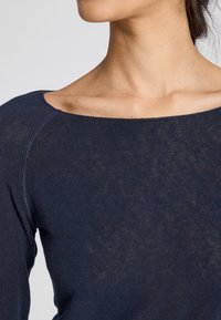 Pulover ajustat de culoare bleumarin, cu un decolteu larg, realizat dintr-un material tricotat ușor, care are o textură subtilă și detalii cusute pe umeri.