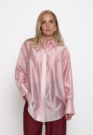 Femme blonde portant une chemise oversize brillante rose clair à boutons et un pantalon rouge foncé, debout devant un fond clair uni.