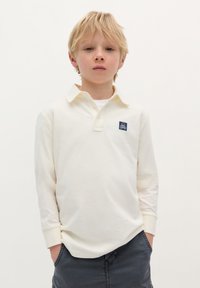 Polo shirt crem cu mâneci lungi și guler, cu un mic petic albastru pe piept. Purtați cu pantaloni scurți gri.