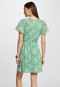 Grün geblümtes Kleid mit tailliertem Schnitt, kurzen schwingenden Ärmeln und weichem Stoff. Verfügt über einen Reißverschluss im Rücken und zarte Muster.