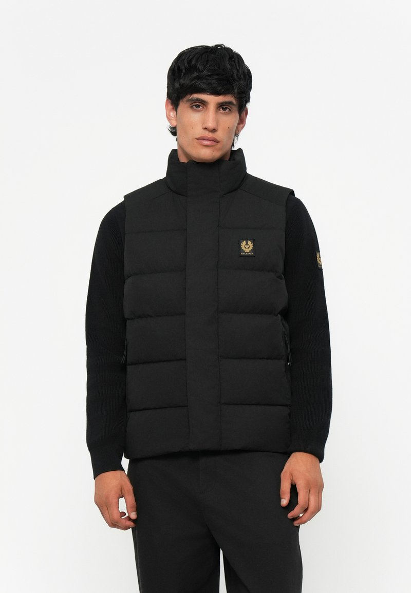 Black padded vest with high collar and golden logo. Features a quilted design, side zippers, and a fitted silhouette. Fabric appears smooth.