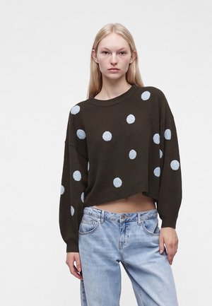 Jeune femme aux longs cheveux blonds porte un pull ample foncé à pois clairs et un jean bleu clair sur un fond uni.
