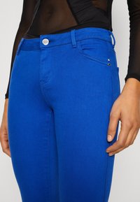 Jeans de mezclilla azul con corte ajustado, que cuentan con un botón plateado, dos bolsillos frontales y costuras decorativas en los laterales. Textura suave.
