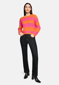 Pull en tricot à rayures orange vif et rose, encolure inclinée, manches longues, porté avec un jean noir à jambes droites et des bottes noires à talons.