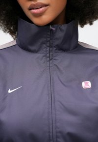 Μωβ ελαφρύ μπουφάν με ψηλό γιακά, λογότυπο Nike και ροζ τετράγωνη πλακέτα. Διαθέτει εμπρόσθιο φερμουάρ και λεία υφή.