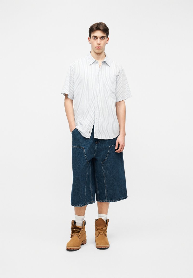 Ung mand står iført en kortærmet hvidt stripede skjorte, brede denim knælange shorts, hvide sokker og brune snørestøvler.
