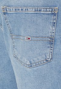 Tela de mezclilla azul con un bolsillo trasero rectangular. El bolsillo presenta un pequeño parche de logo en colores rojo, blanco y azul.