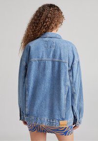 Ljusblå denimjacka med en klassisk passform, prydd med en framficka och en tagg som visar varumärket, bärs över en mönstrad kjol.