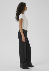Wide-leg black denim jeans with a raw hem, paired with a fitted white short-sleeve shirt. Minimalist design with straight silhouette.