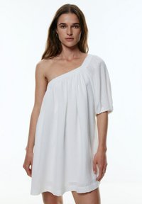 Robe blanche à une épaule fabriquée dans un tissu léger. Elle présente une coupe ample, une surface texturée et une manche bouffante sur la droite.