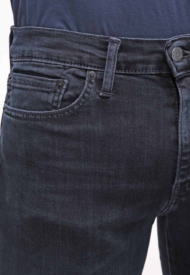 Jean en denim bleu foncé à coupe ajustée, avec une fermeture à boutons, des poches avant, et une surface texturée avec des détails de coutures subtiles.