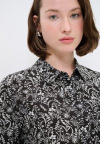 Maglietta nera con stampa floreale bianca, design con colletto, tessuto leggero e dettagli raffinati nei motivi floreali.