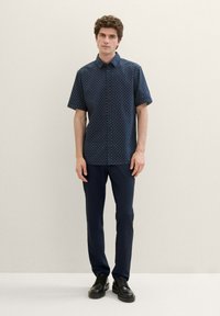 Navy short-sleeve shirt with a geometric print, button-up design, paired with dark pants and black shoes. Texture appears smooth and breathable.