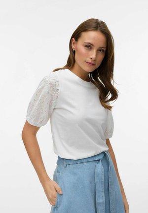 Vrouw draagt een witte blouse met pofmouwen en eyeletdetails en een lichtblauwe, hooggetailleerde spijkerrok met een gestrikte riem, poserend met één hand in de zak.