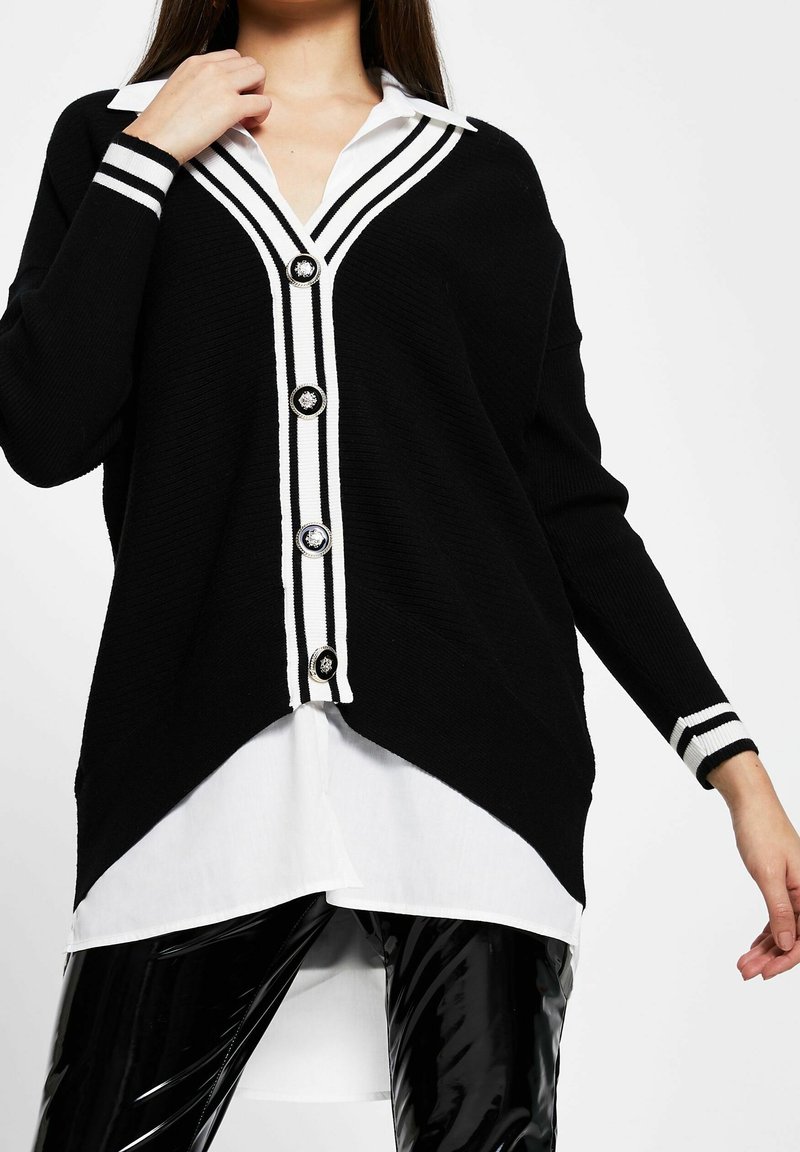 Zwarte gebreide cardigan met witte gestreepte accenten, voorzien van een V-hals, knoopsluiting en een asymmetrische zoom gedragen over een wit shirt.