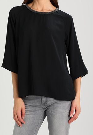 Woman wearing a black loose-fit blouse with three-quarter sleeves and gray jeans, standing against a plain background.