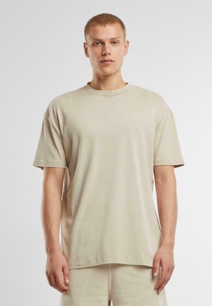 Jeune homme aux cheveux courts portant un T-shirt beige ample et un short beige, se tenant devant un fond blanc uni.