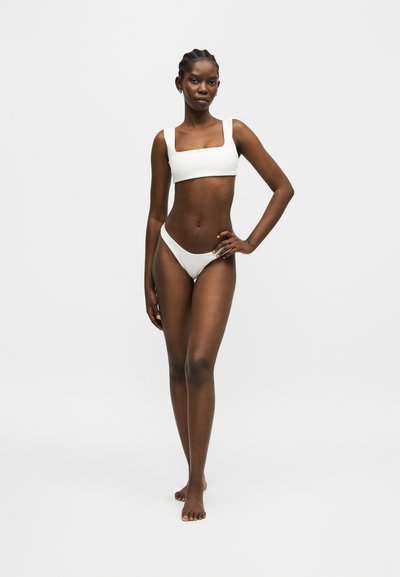 Γυναίκα με πλεξούδες που φοράει λευκό μπικίνι με τετράγωνο ντεκολτέ, στέκεται ξυπόλυτη με το ένα χέρι στη μέση σε απλό φωτεινό φόντο.