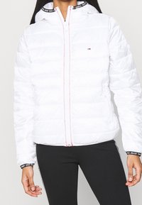 Giacca a vento bianca con chiusura a zip, dotata di cappuccio e polsini accentuati in nero e rosso, texture trapuntata e dettagli in cucitura rossa.
