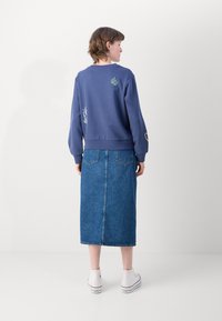 Sweatshirt violet avec des accents brodés, associé à une jupe midi en denim. Des chaussures blanches à enfiler complètent la tenue. Vue de dos.