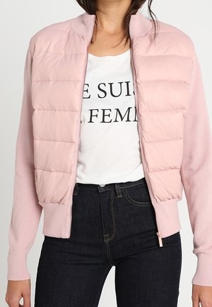 Woman wearing light pink quilted bomber jacket over white shirt with "JE SUIS FEMME" text, paired with high-waisted dark jeans.
