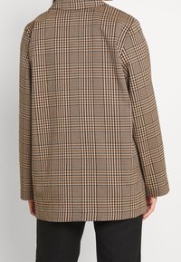 Hnědo-beige károvaná bunda s volným střihem, s límcem a dlouhými rukávy, vyrobená z texturované látky s polodlouhým designem.