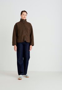 Brown fleece jacket with a high collar, front zip, and two pockets, paired with dark denim wide-leg pants and silver sneakers.