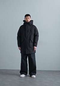 Schwarze Parka mit hohem Kragen und verstellbarem Kapuze, ausgestattet mit zwei Seitentaschen und einer geraden Silhouette, kombiniert mit weiten schwarzen Hosen.