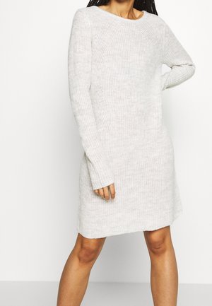 Frau trägt ein langärmliges, hellgraues gestricktes Pulloverkleid und steht mit einer Hand in der Hüfte vor einem schlichten Hintergrund.