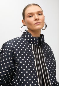 Námořnický puffer kabát s puntíky, vysokým límcem, předním zipem a černými akcenty. Obsahuje velké stříbrné kruhové náušnice.