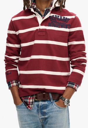 Mann trägt ein weinrot-weiß gestreiftes Rugbyshirt über einem rot karierten Hemd, mit blauer Jeans, braunem Gürtel und schwarzer Armbanduhr.